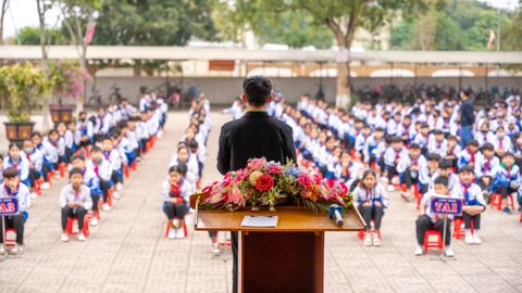 A teacher addresses students seated in an outdoor assembly at a school.