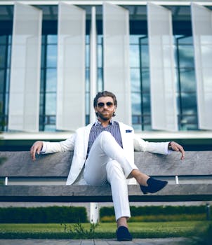 一位身穿白色西装的时尚男士自信地坐在户外长凳上，展现出优雅与现代的生活方式