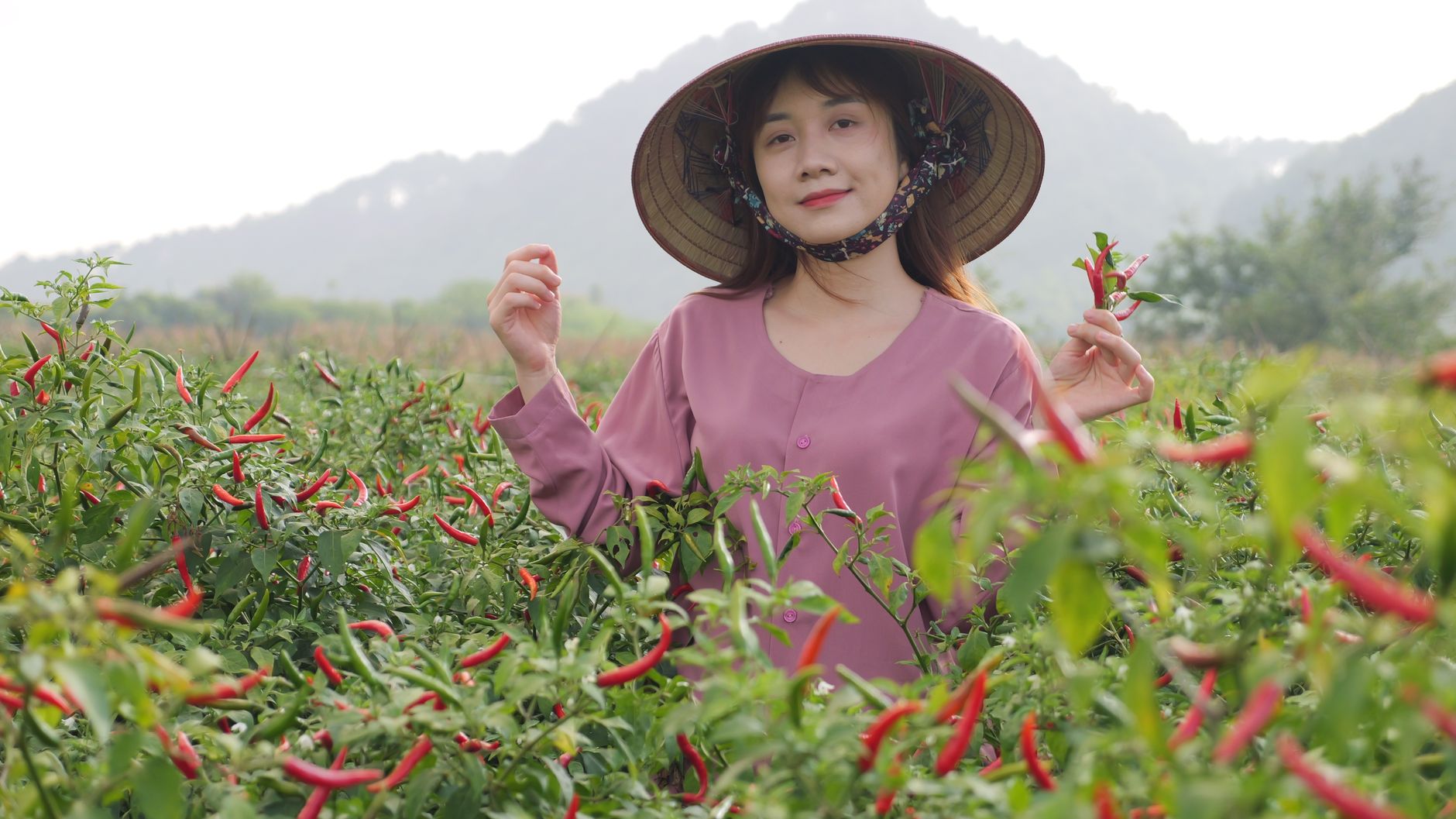 A smiling Asian woman wearing a hat is standing in a rural field with red peppers, showcasing traditional agriculture.