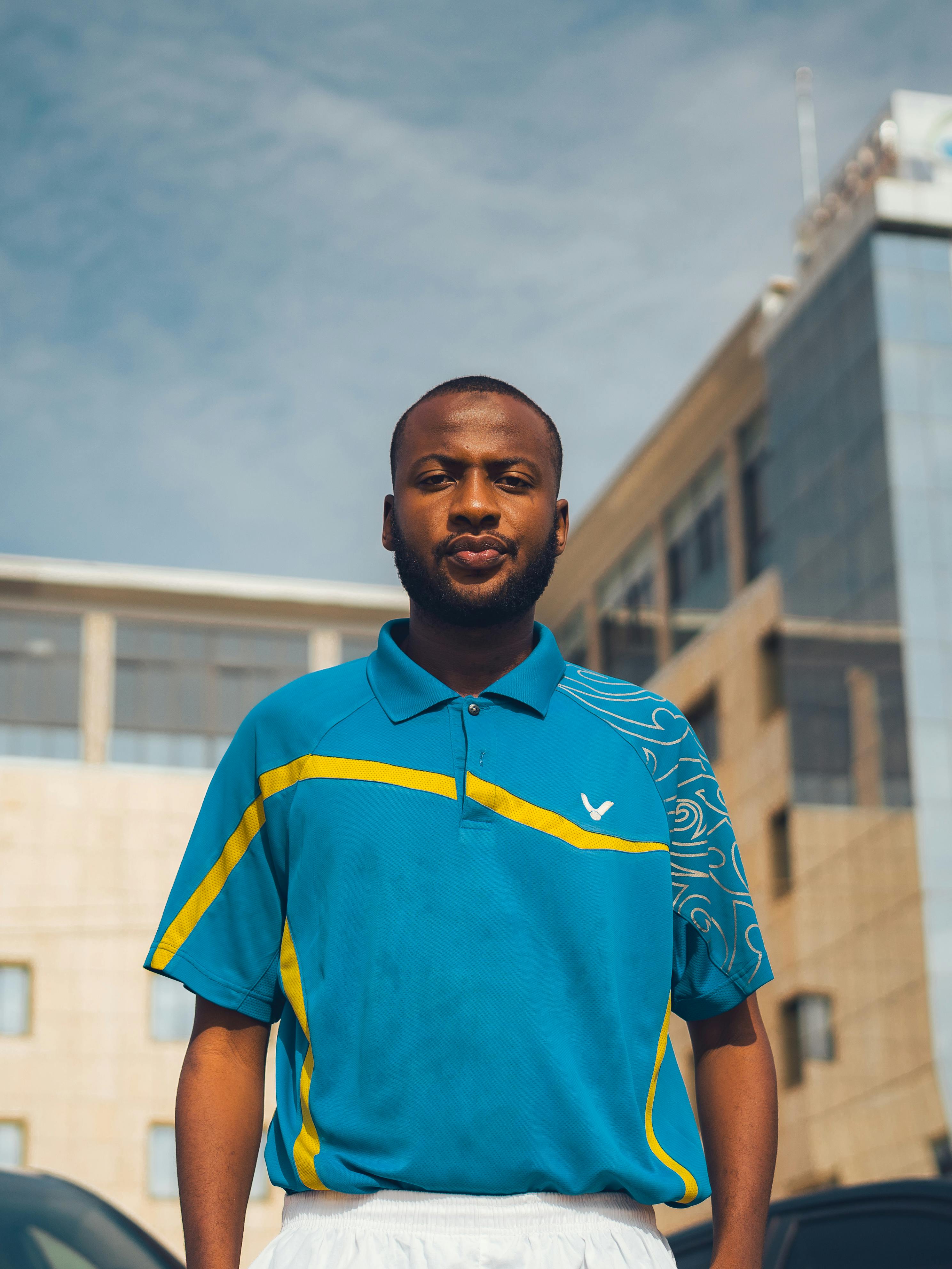 A confident young man in a blue polo shirt stands outdoors in Abuja, Nigeria, with an office building behind.
