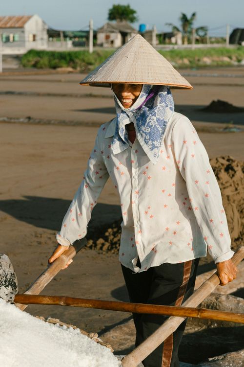 Free A cheerful farmer wearing a conical hat pushes a wheelbarrow filled with salt at a rural field in Vietnam. Stock Photo