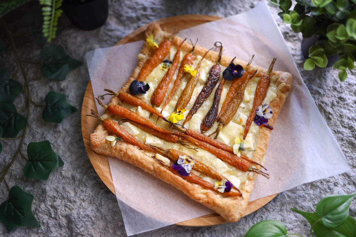 Rectangular puff tart topped with whole orange and purple carrots, cheese and edible flowers