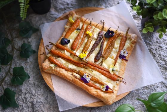 Rectangular puff tart topped with whole orange and purple carrots, cheese and edible flowers
