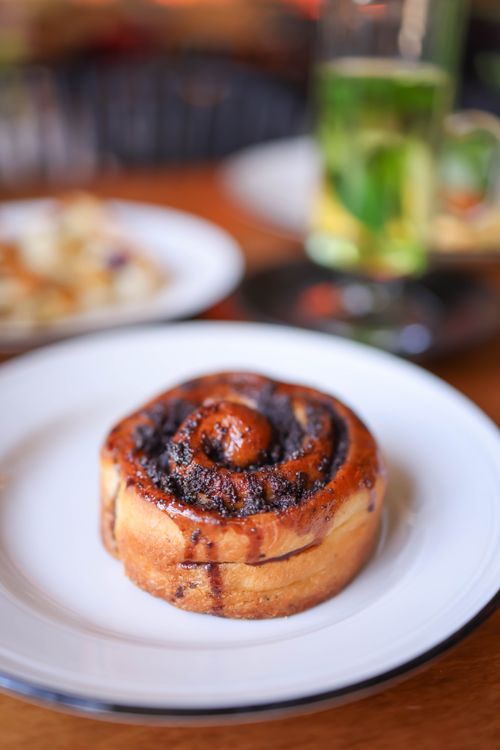 Free Appetizing cinnamon roll on a white plate with blurred background of tea. Perfect for breakfast themes. Stock Photo