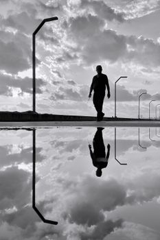 Silhouetted man walking on a road with reflection, dramatic clouds.