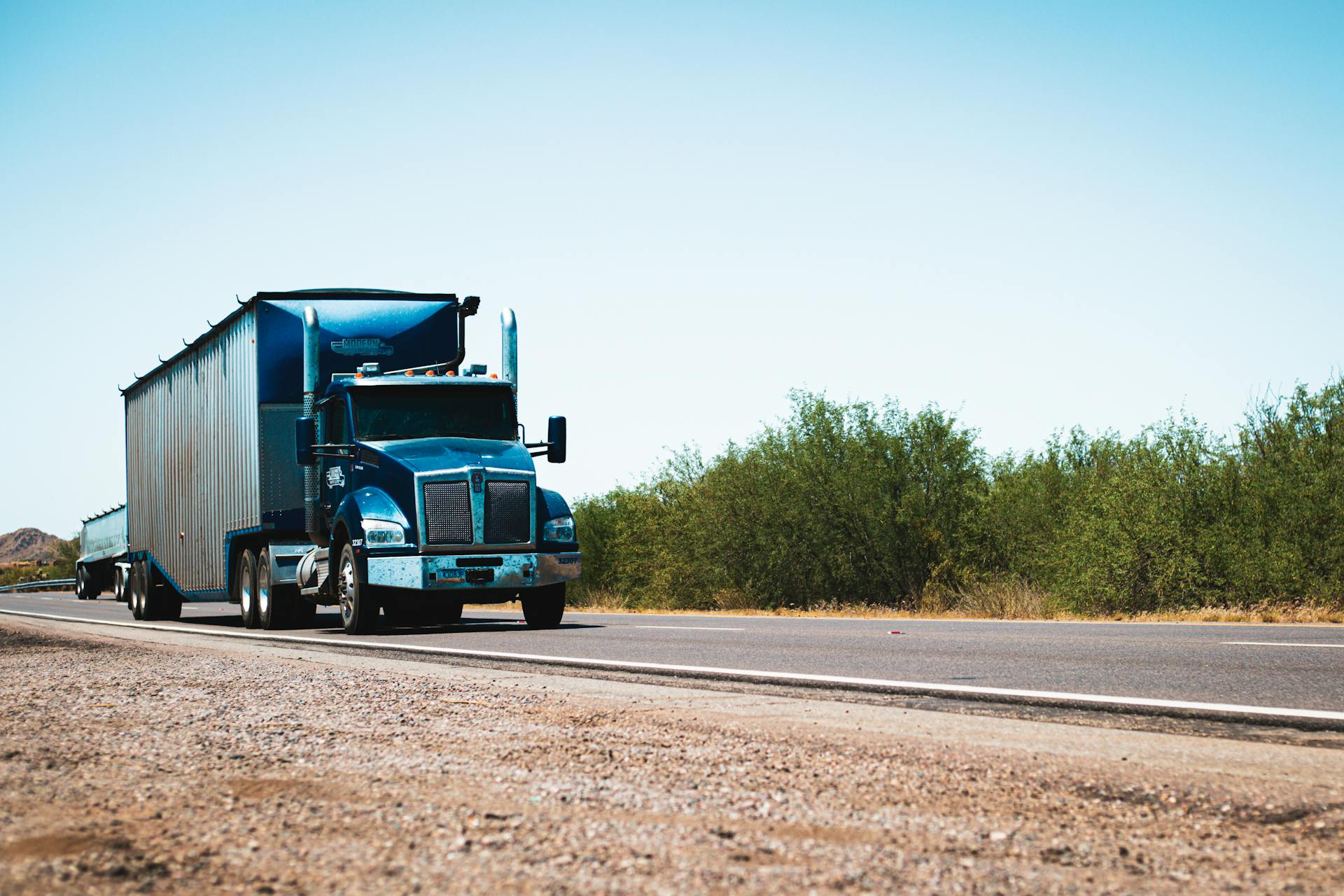 A blue semi truck driving on a rural highway under clear blue skies, showcasing transportation and travel.