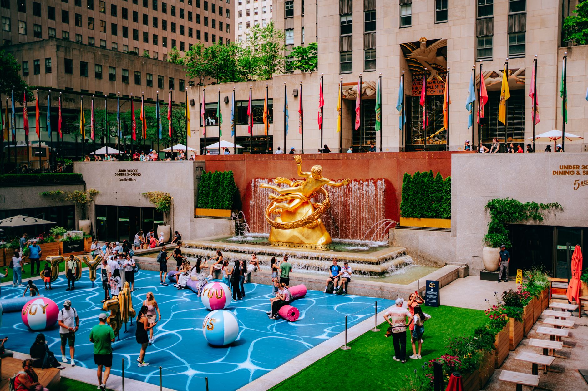 Vibrant scene at Rockefeller Center with the iconic Prometheus statue, flags, and playful activities.