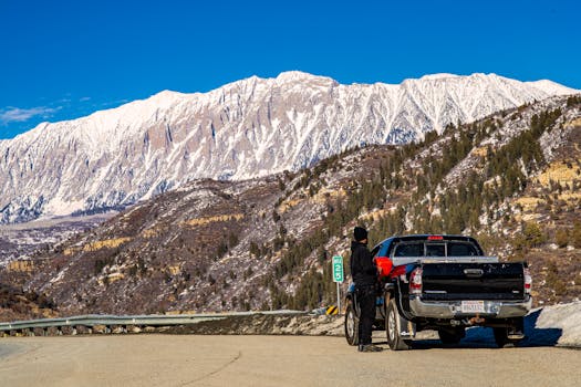 A man stands by a pickup truck enjoying the stunning view of snow-covered mountains on a clear day.