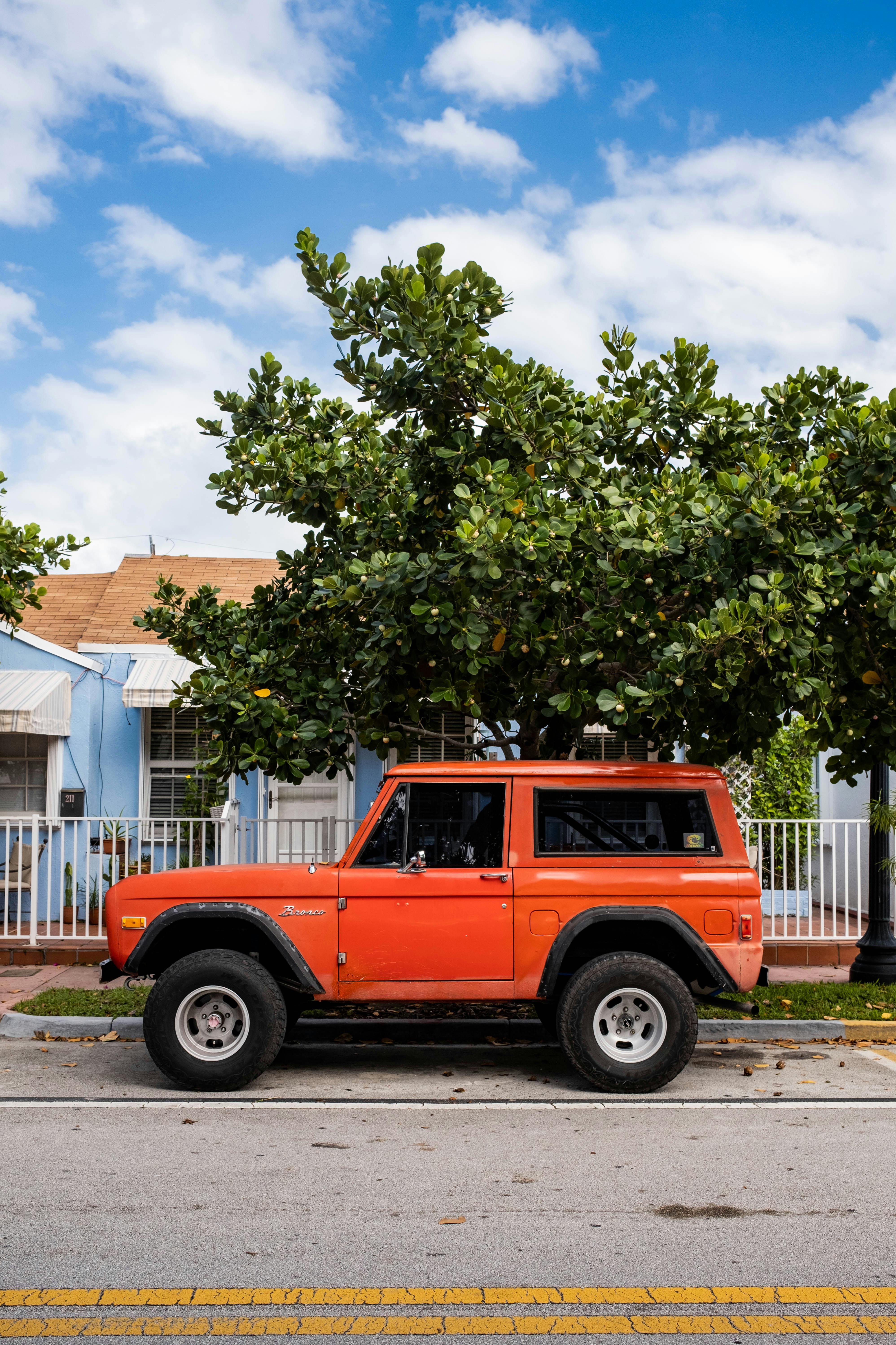 A vintage orange SUV parked on a street by a vibrant blue house with lush greenery, under a clear blue sky.
