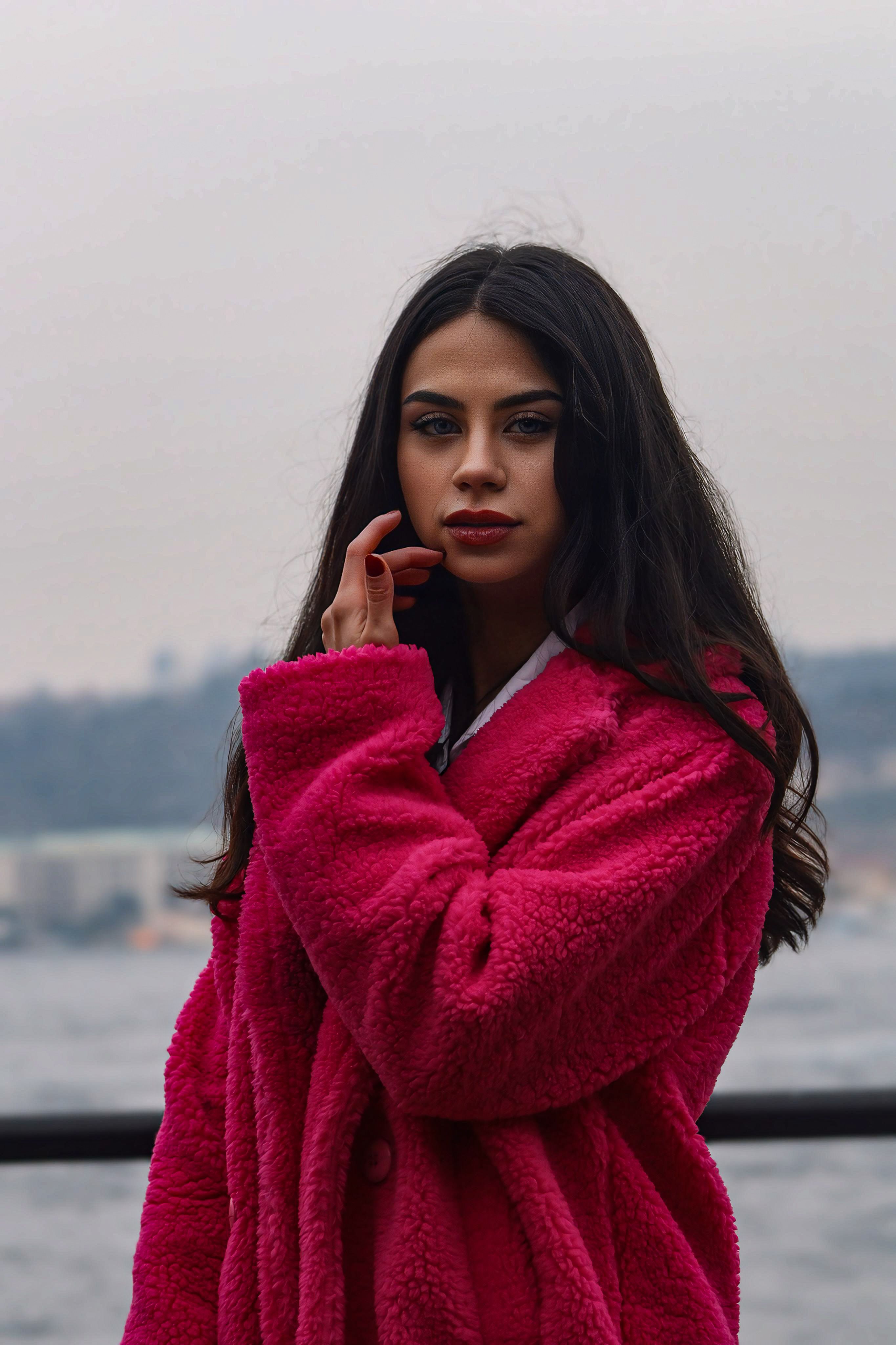 Fashionable woman in a vibrant pink coat posing outdoors by the water.