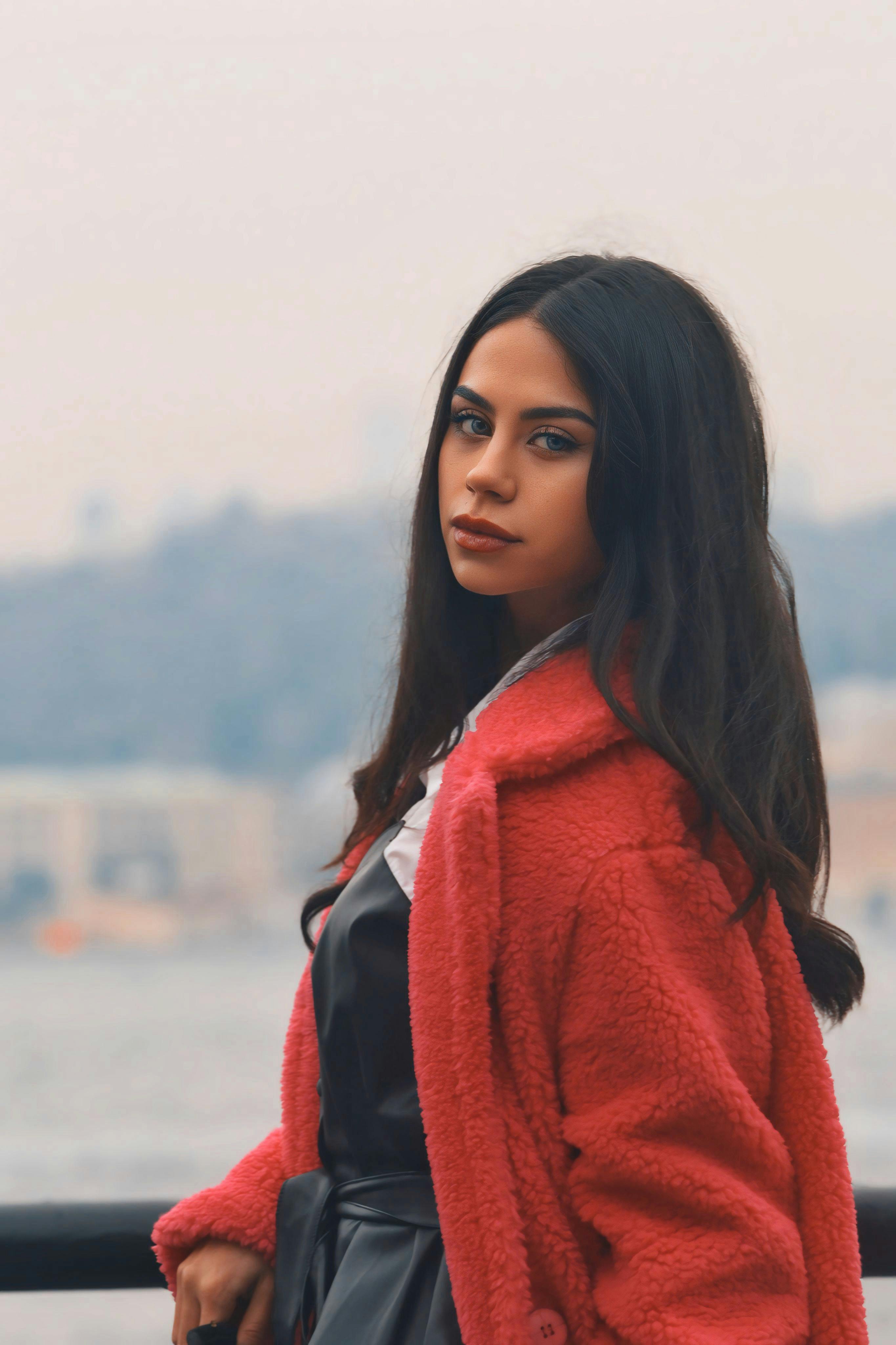 Elegant woman in a red coat poses near a scenic water backdrop, evoking serene and stylish vibes.