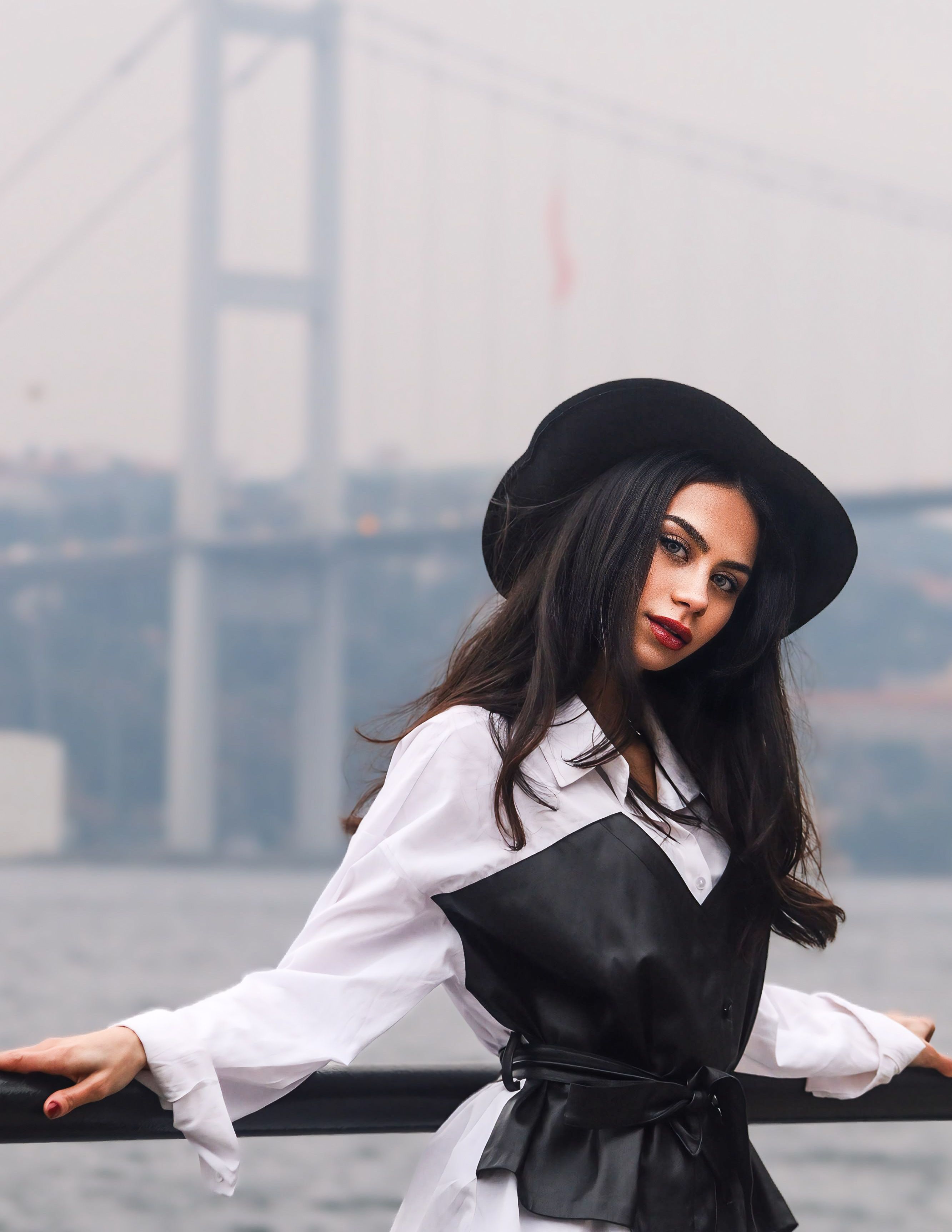 Fashionable woman in black hat and outfit posing by a famous bridge for a modern portrait.