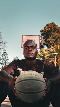 A young adult male sitting on an outdoor court holding a basketball with a hoop in the background.