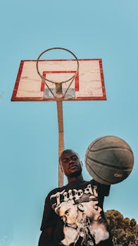 A young man poses with a basketball beneath a hoop against a clear blue sky, capturing sports energy.