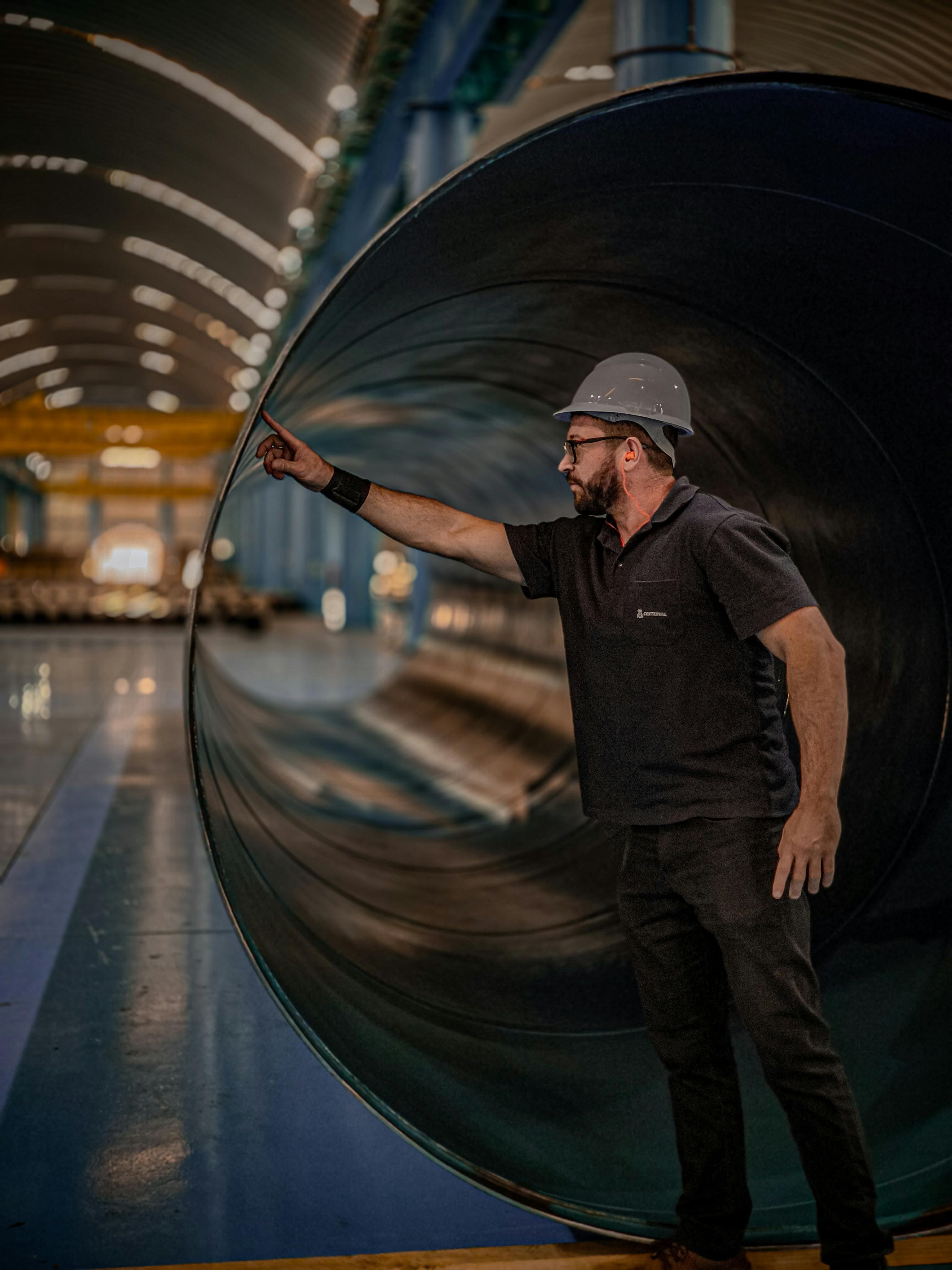 Engineer in helmet assessing a large pipe in a manufacturing warehouse, showcasing industrial productivity.