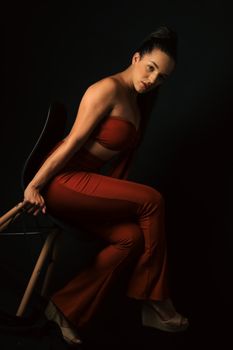 A fashionable woman in elegant attire poses on a chair in a dark studio. Glamorous and sophisticated portrait.
