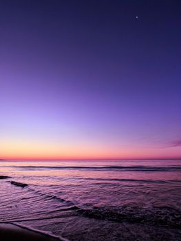 A tranquil ocean scene at sunset with vibrant purple hues and gentle waves under a crescent moon.