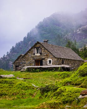 Rustic stone house nestled in lush Swiss Alps, surrounded by greenery and misty mountains.