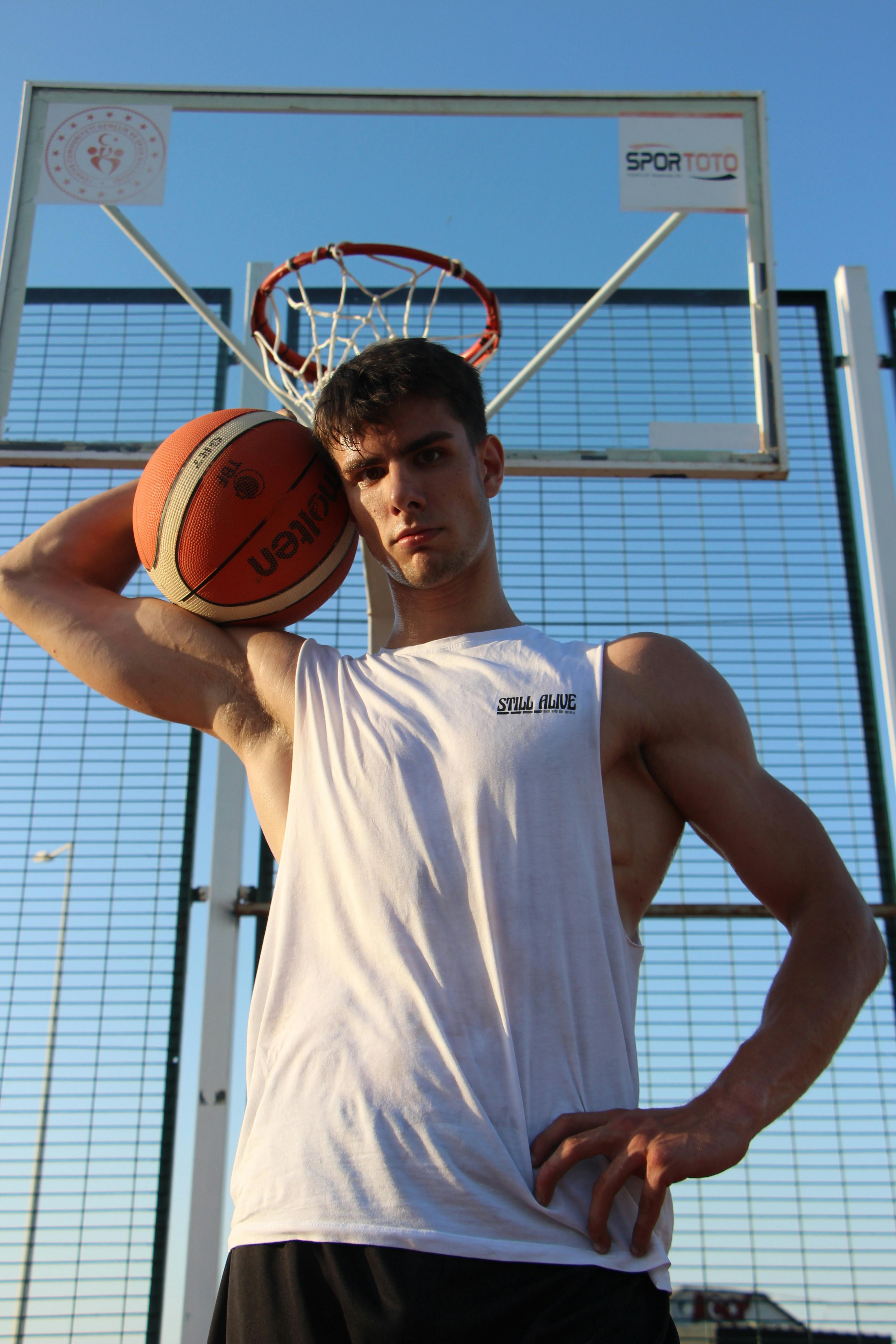 Caucasian man in a tank top poses with a basketball on an outdoor court in Türkiye.