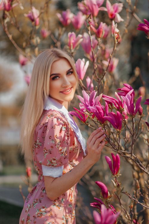 Free Charming blonde woman in a pink floral dress posing with vibrant magnolia blossoms in a sunny garden. Stock Photo