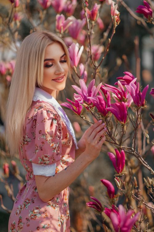 Free Blonde woman in floral dress admiring pink magnolia flowers outdoors on a sunny day. Stock Photo