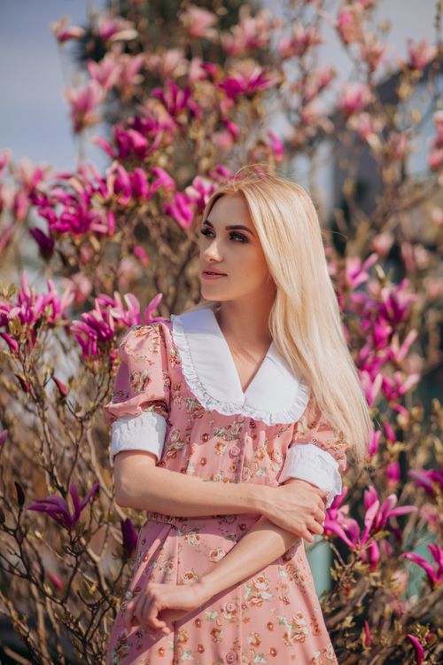 Free A young blonde woman in a vintage pink floral dress stands among blooming magnolia trees in a sunlit park. Stock Photo