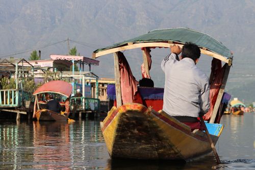 Free A man rowing a wooden boat on a serene lake surrounded by colorful structures and distant mountains. Stock Photo