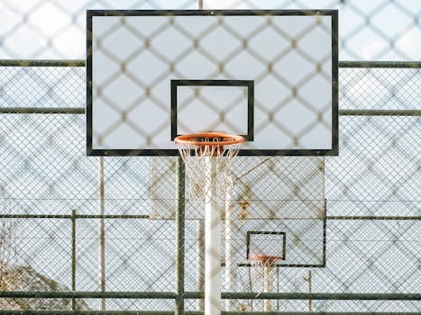 A basketball hoop seen through a chainlink fence on an outdoor court under daylight.