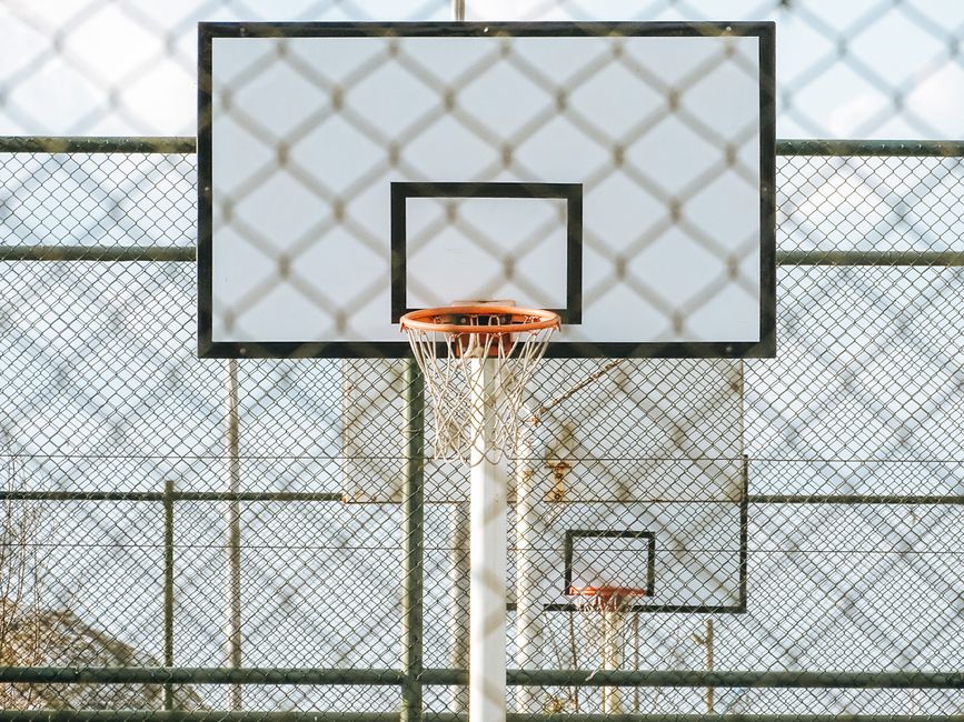 A basketball hoop seen through a chainlink fence on an outdoor court under daylight.