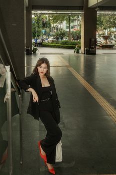 Elegant woman in black outfit poses stylishly by a railing in a modern urban environment.