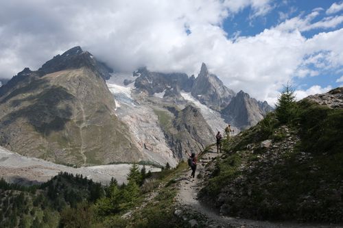 Free Hikers explore a scenic mountain trail with dramatic views of rocky peaks and lush greenery under a cloudy sky. Stock Photo