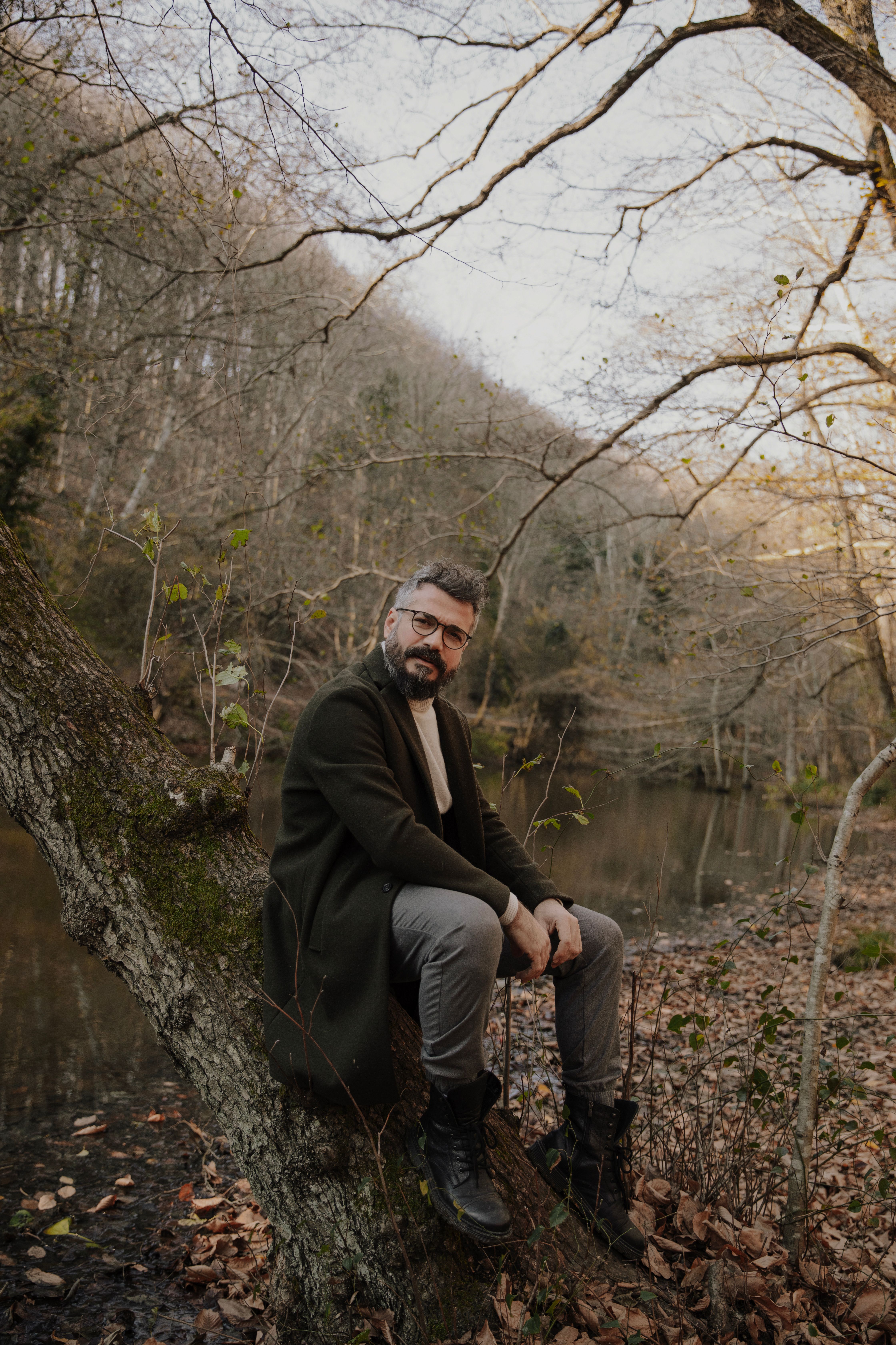 A fashionable man in a coat sits on a tree by a forest pond in autumn.