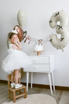 A young girl in a white dress celebrates her third birthday with balloons and cake.