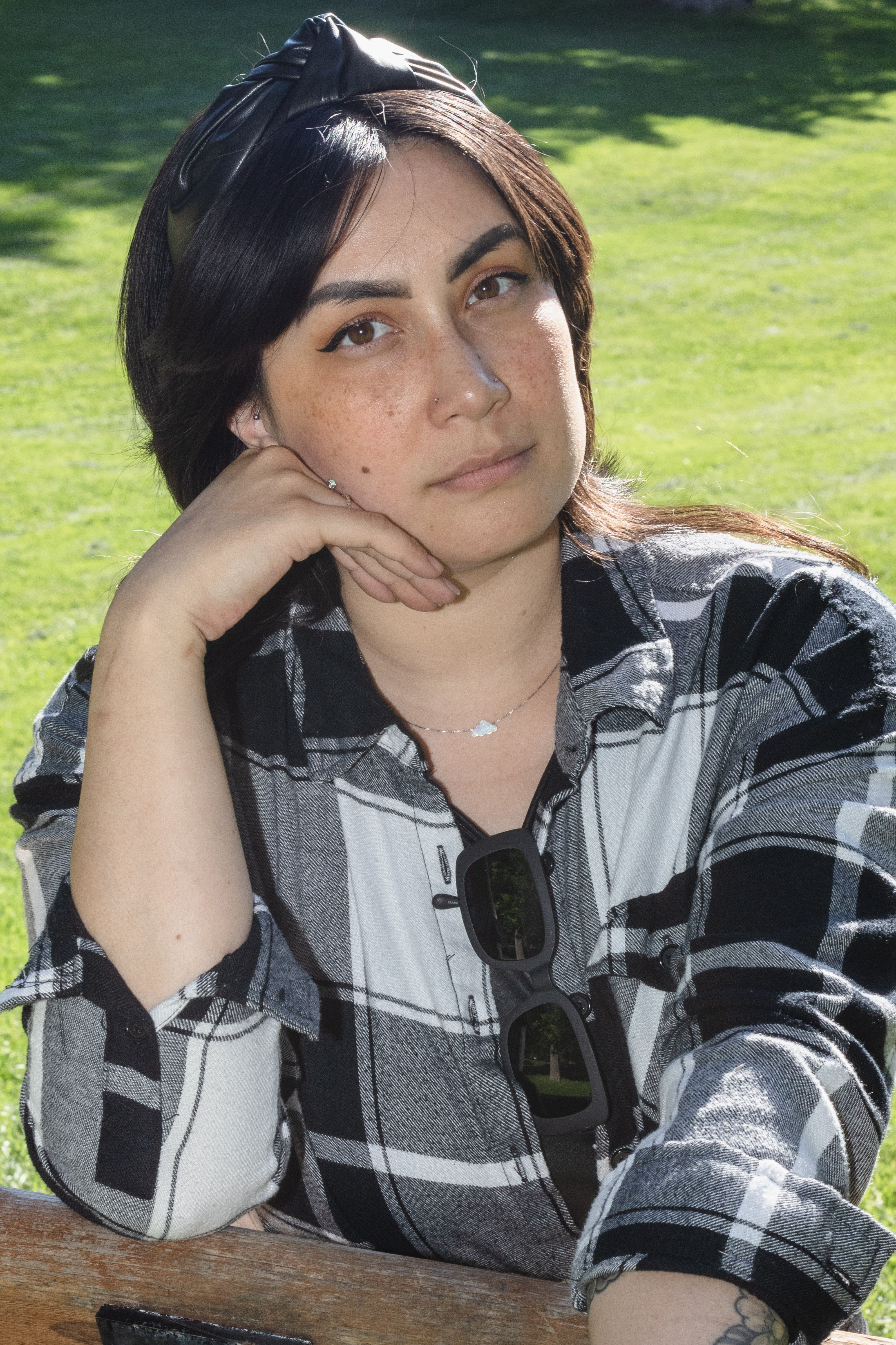 A thoughtful woman sitting outdoors in a checkered shirt, resting her head on her hand with a calm expression.