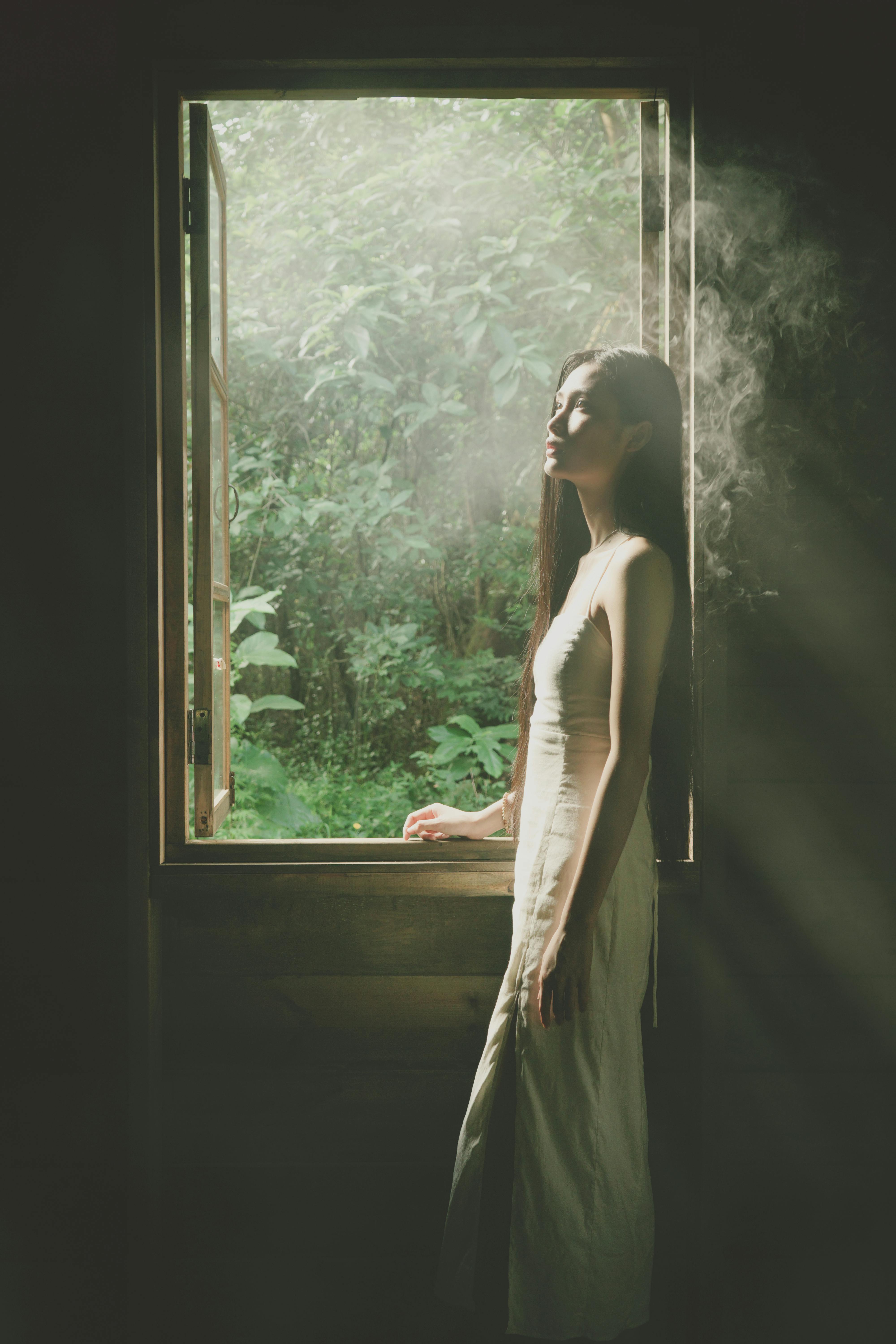 A woman in a white dress enjoys sunlight streaming through an open window with lush greenery outside.