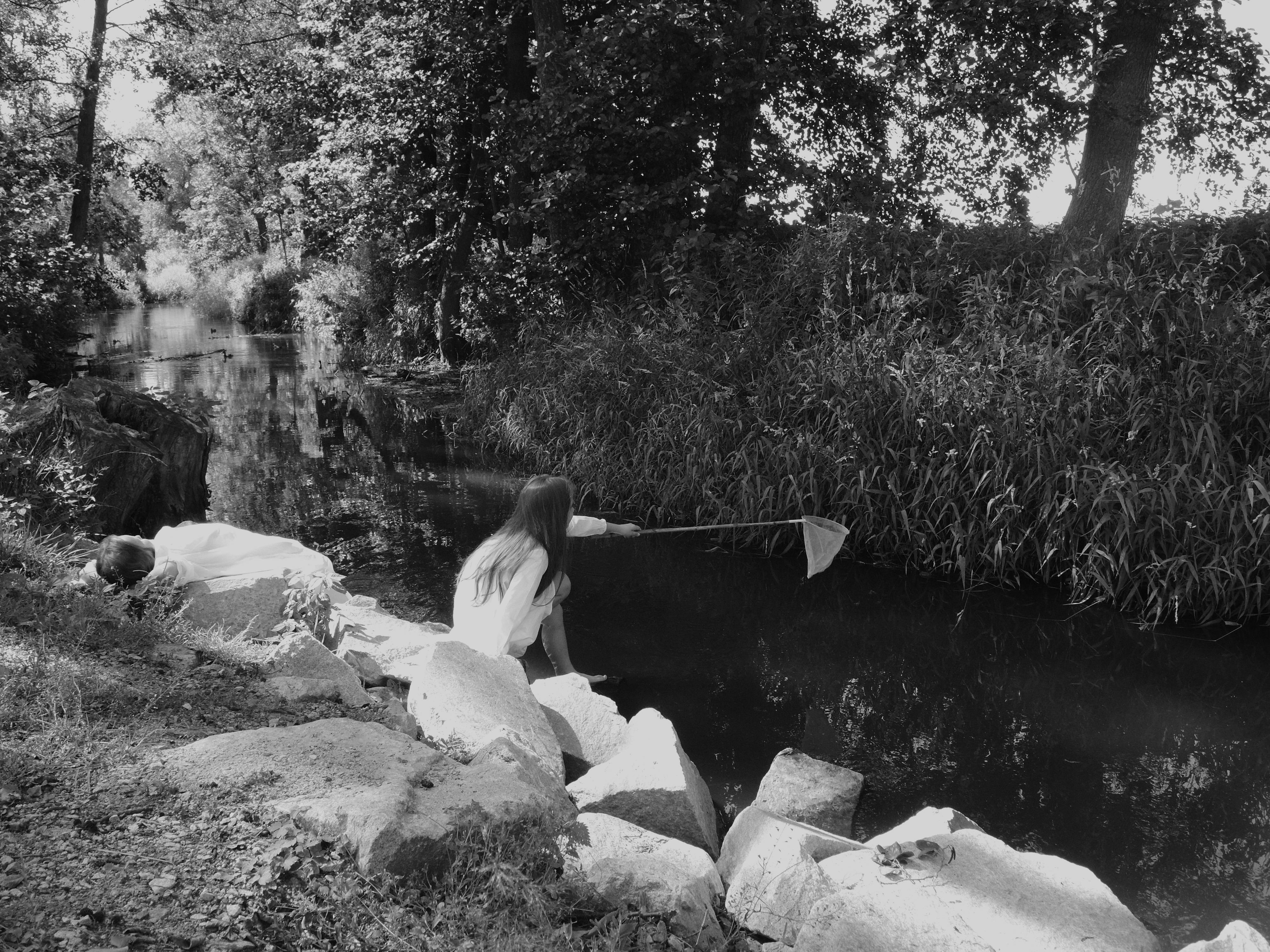 A tranquil black and white image of a woman sitting by a river using a net, surrounded by nature and trees.