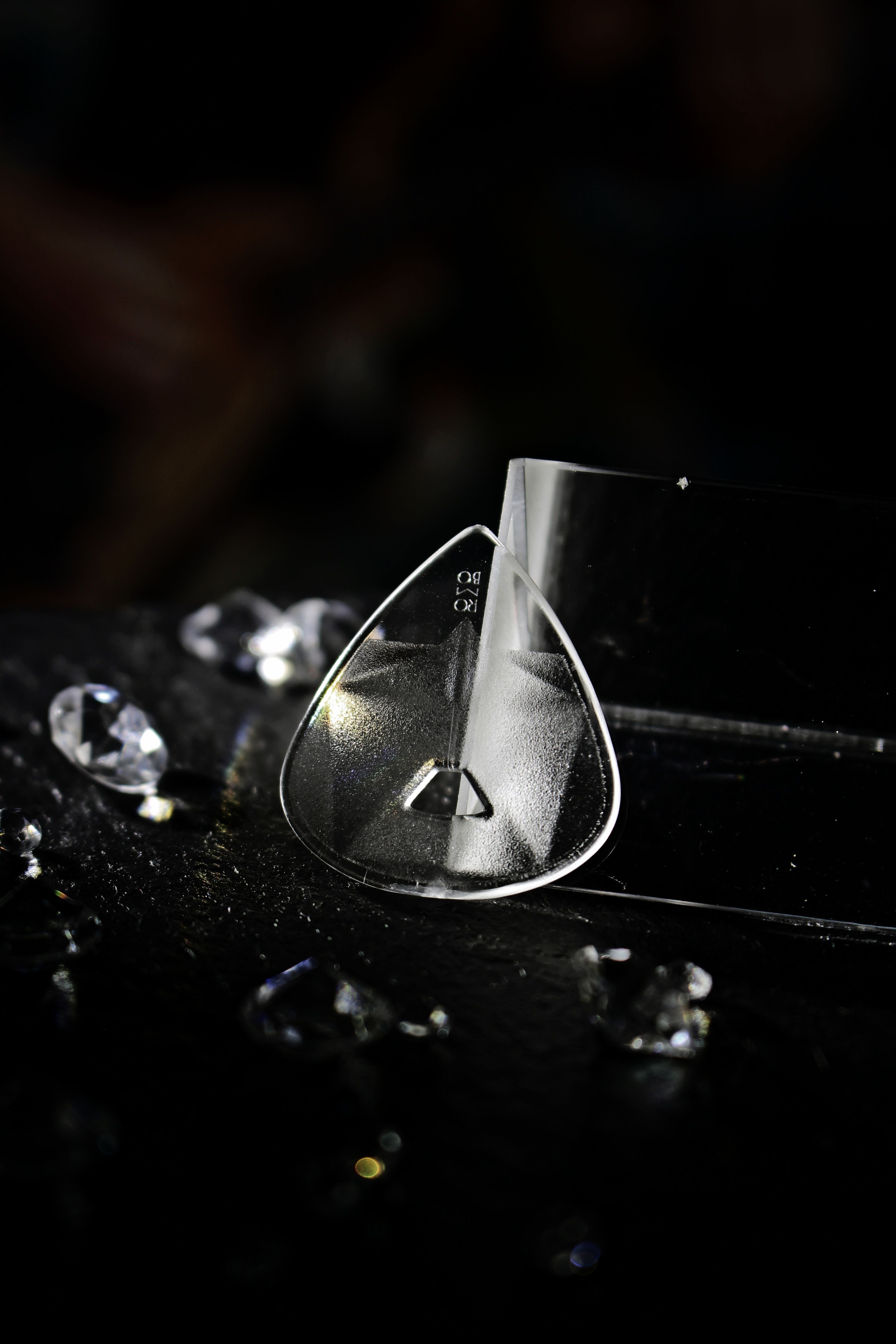 Close-up of a transparent crystal guitar pick surrounded by scattered crystals on a dark background.