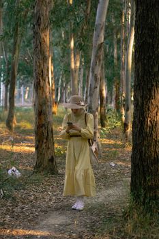 Woman in a yellow dress using a smartphone while walking through a forest path.