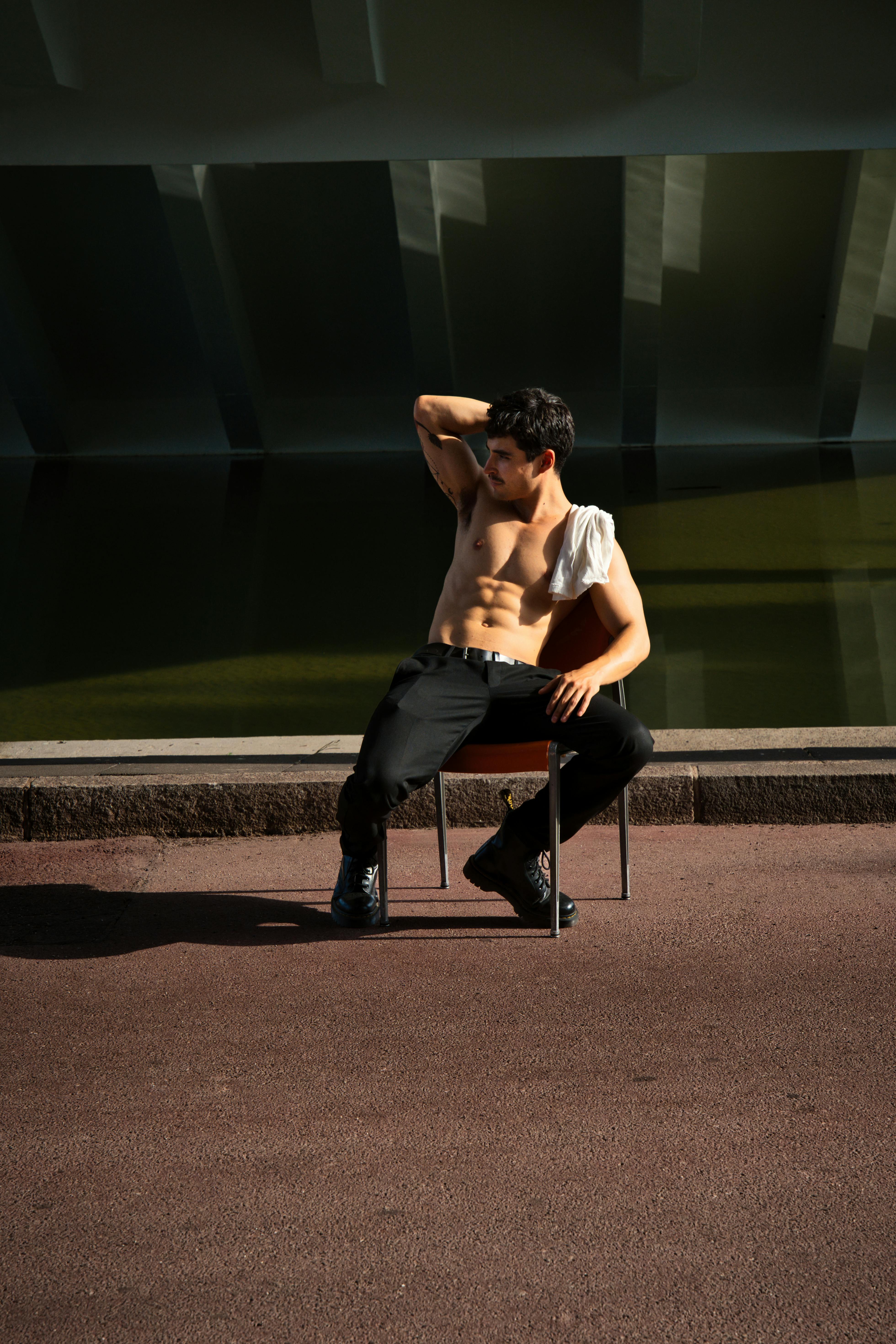 A shirtless man poses confidently while sitting on a chair outdoors in Berlin, Germany.