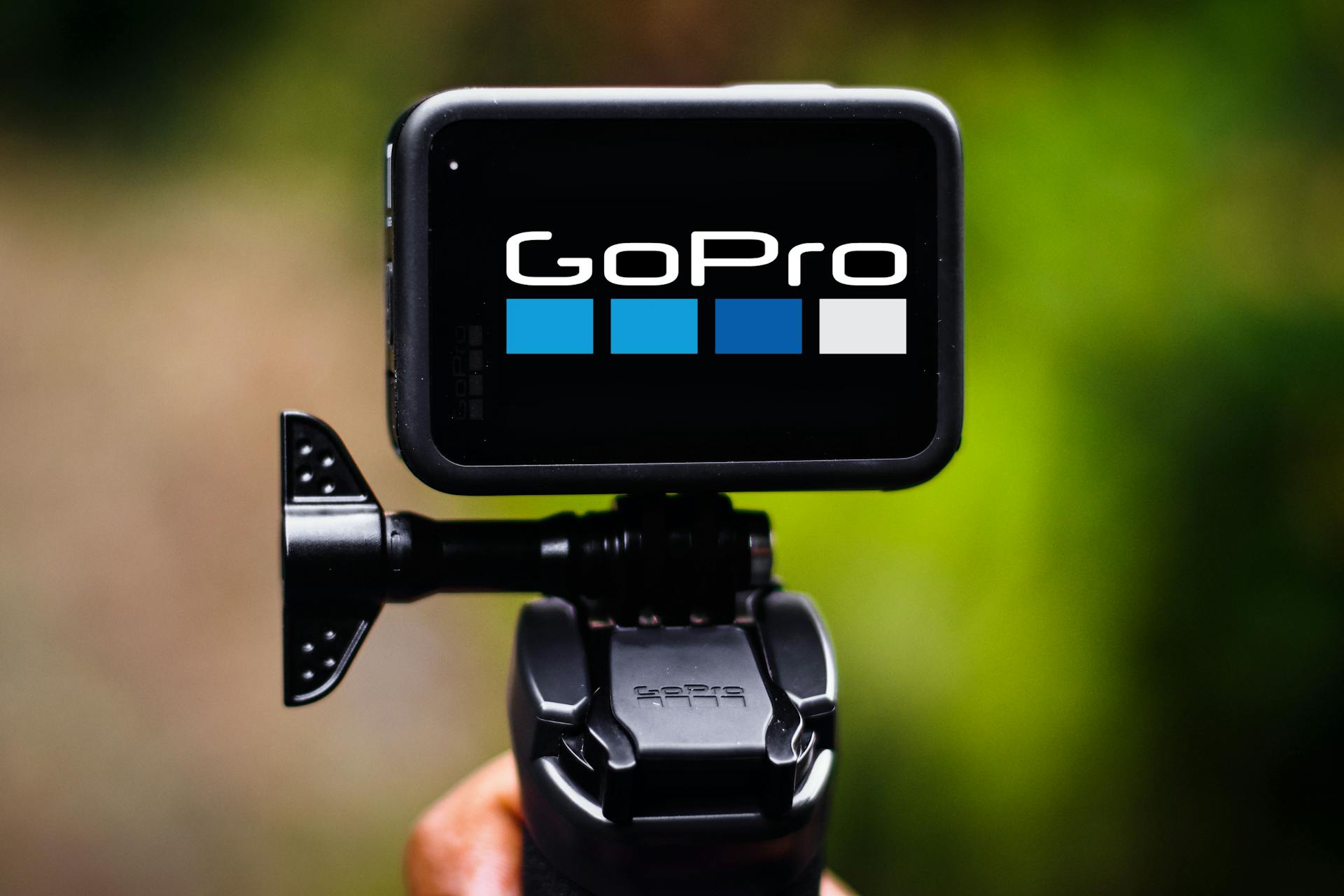 Hand holding a compact action camera on a mount with blurred greenery in the background.