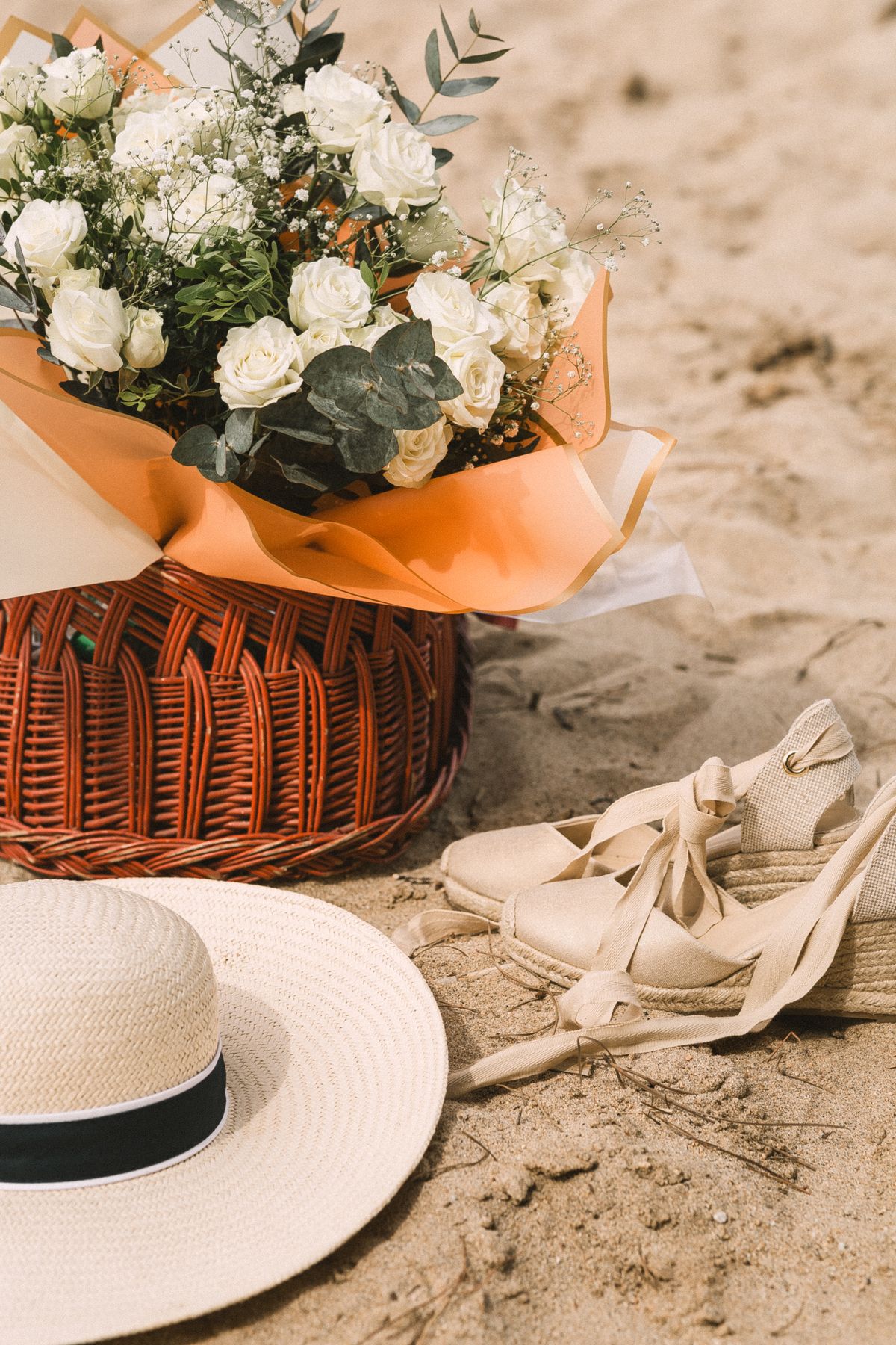 A still life — roses, a basket, a straw hat, and white sandals on the beach
