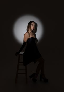 Elegant woman in black dress posing on stool, studio spotlight.