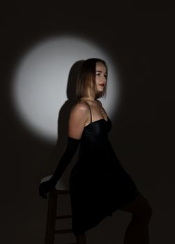 Portrait of a woman in a black dress sitting on a stool under spotlight, creating a dramatic silhouette.