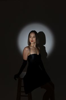 Fashion portrait of a woman in a black dress seated on a stool with dramatic lighting.