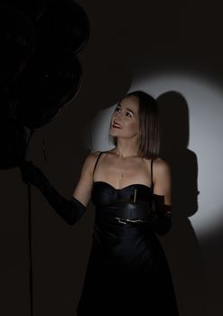 Charming young woman in a black dress holding balloons with dramatic lighting.