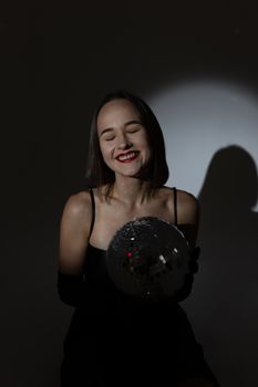 A joyful brunette woman in a black dress holding a disco ball under a spotlight, creating dramatic shadows.
