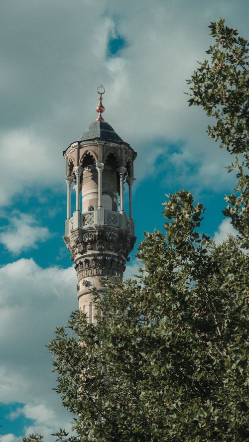 Free A beautifully crafted minaret framed by lush green trees under a vibrant blue sky. Stock Photo