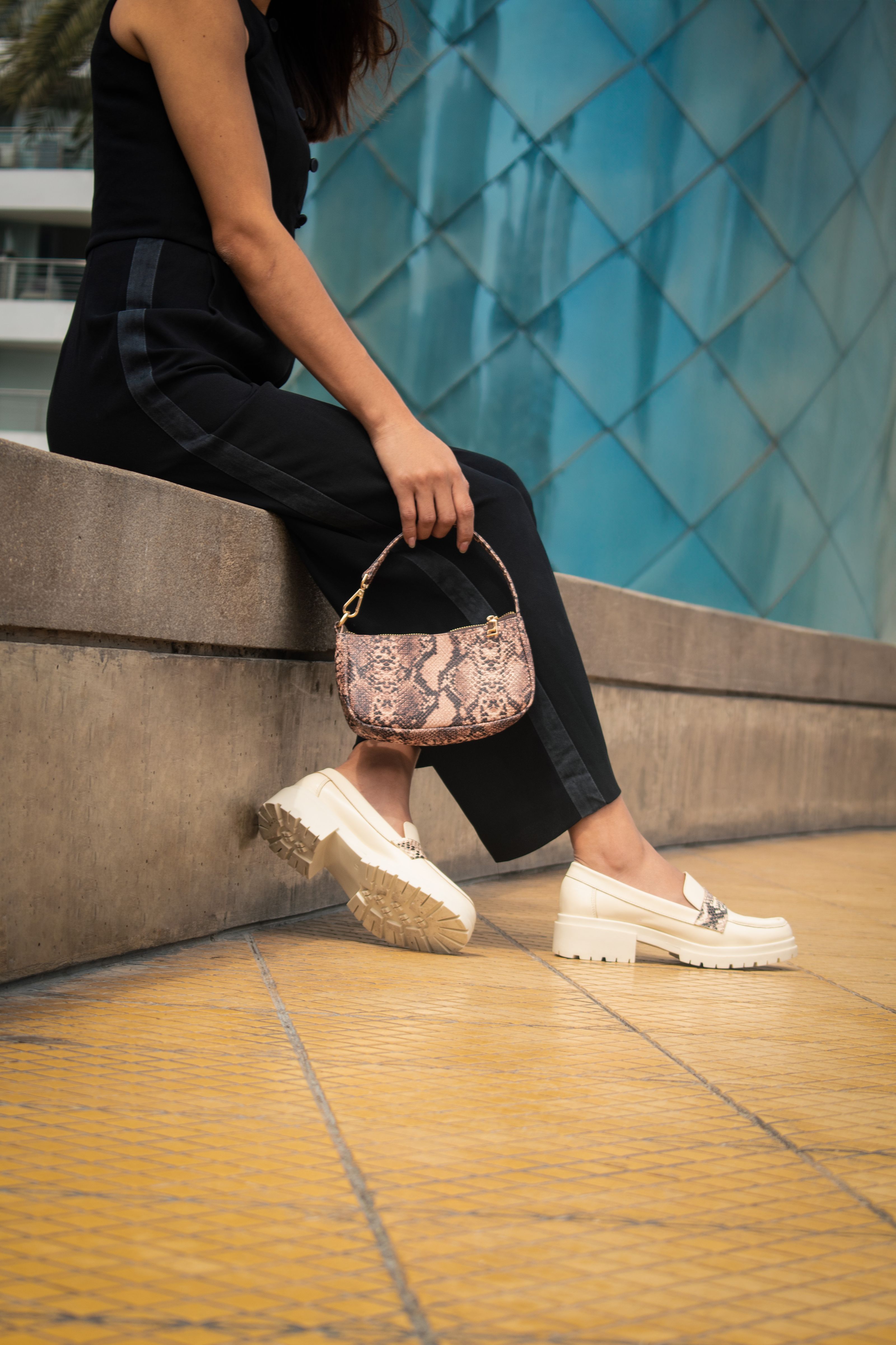 Mulher estilosa posando com bolsa de estampa de cobra e sapatilhas brancas em rua urbana.