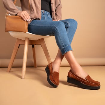 A fashionable woman in jeans and loafers sitting in a studio with leather bag and jacket.