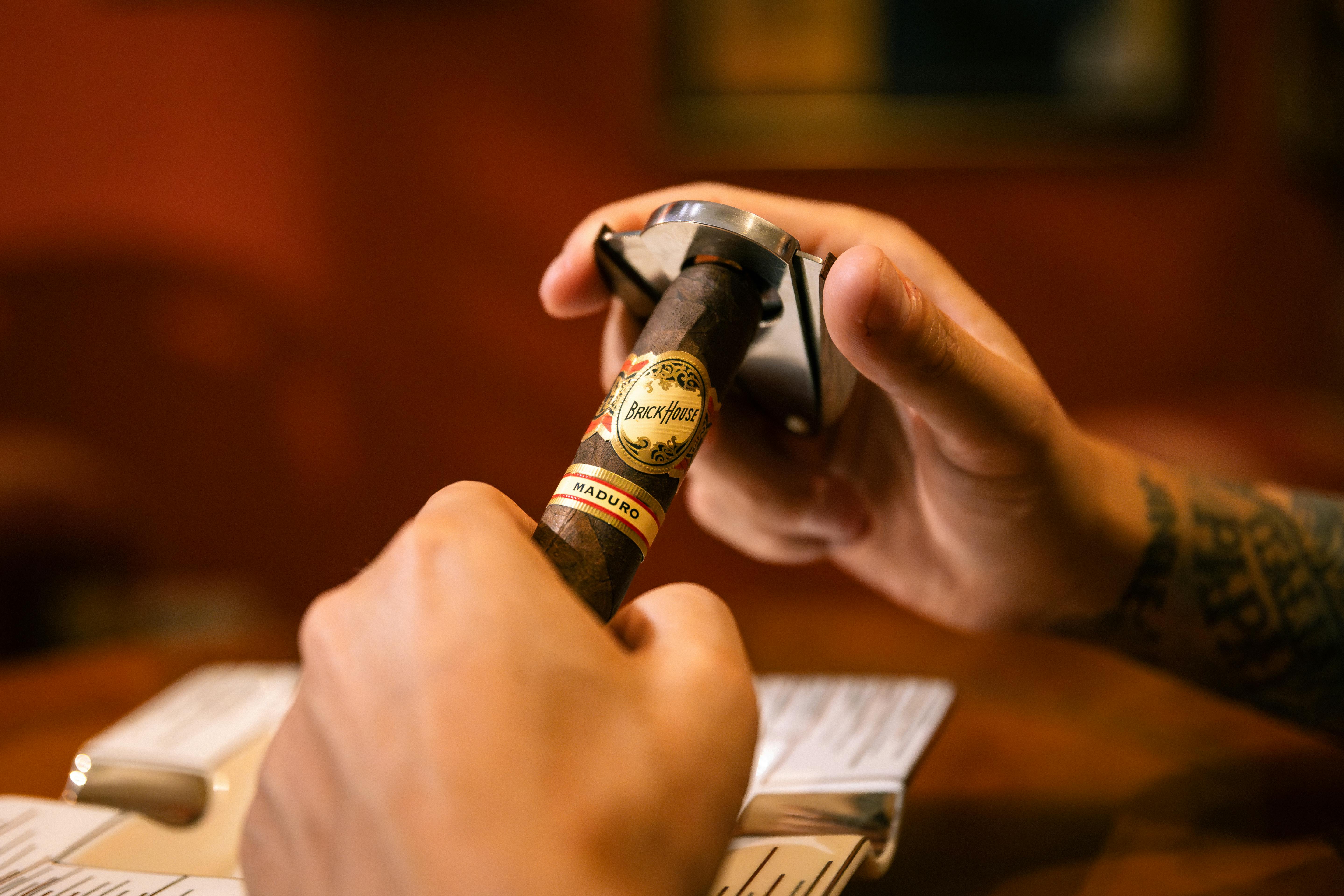 A close-up of hands using a cigar cutter on a BrickHouse Maduro cigar indoors.